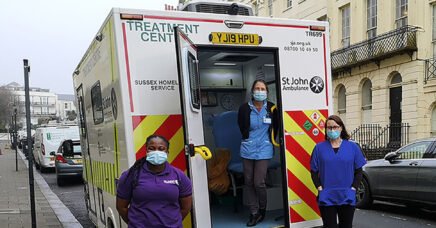 Three people in surgical masks, standing on a city street at the back of an ambulance