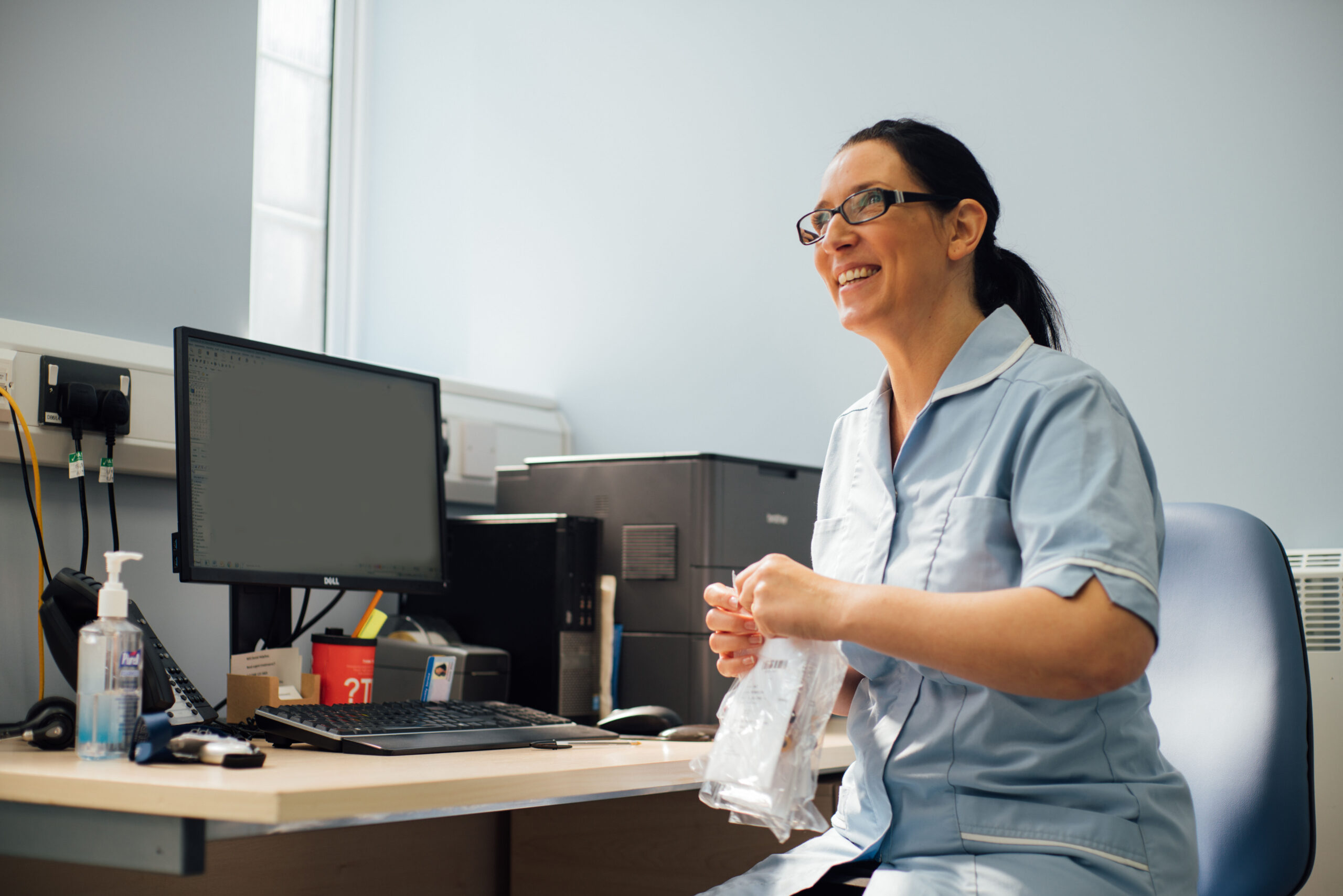 Arch healthcare assistant sits at her desk, smiling at someone off camera
