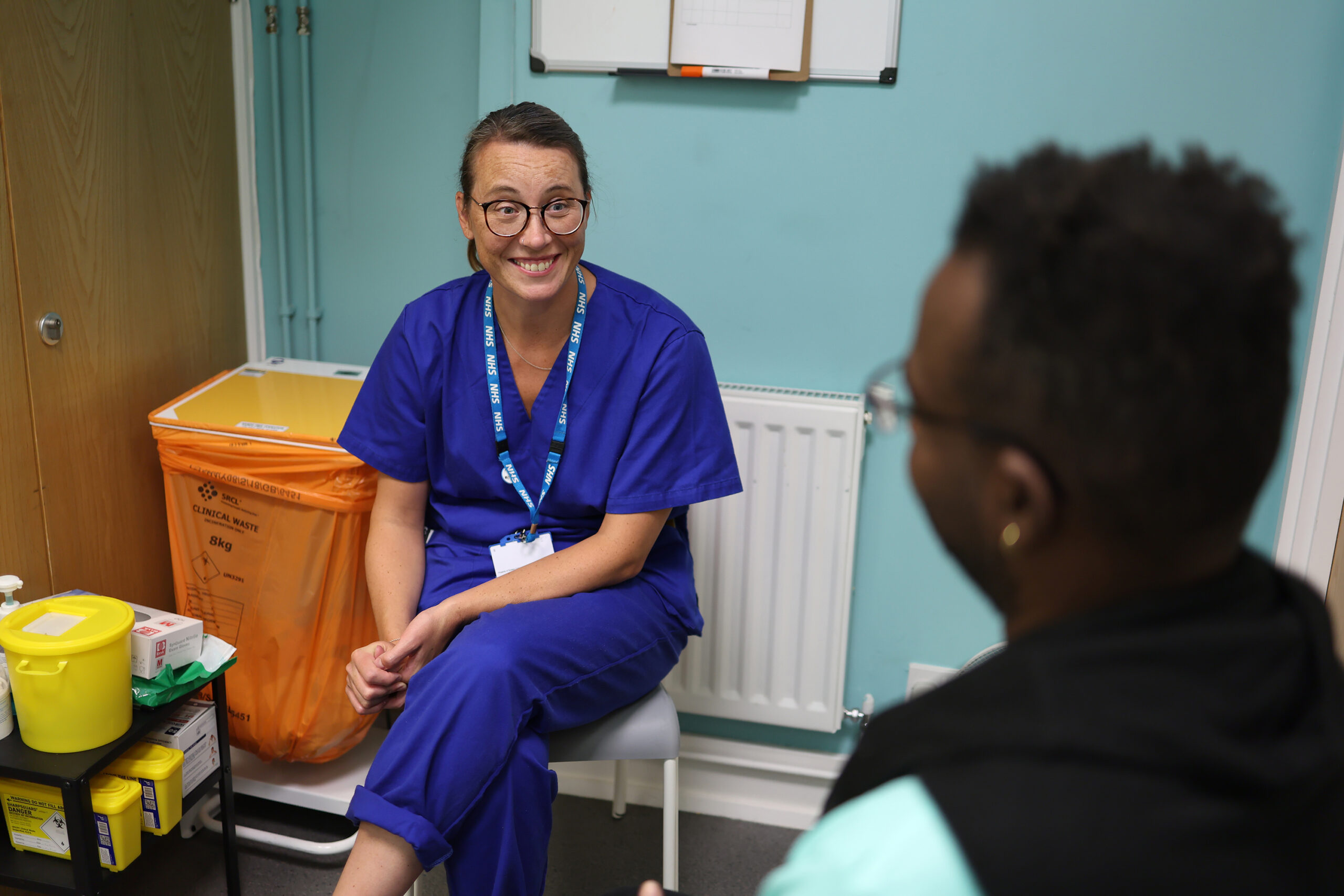A nurse in scrubs sitting in a clinic with a patient, smiling and talking to the patient (back view, blurred), nurse is in focus and smiling at the patient.