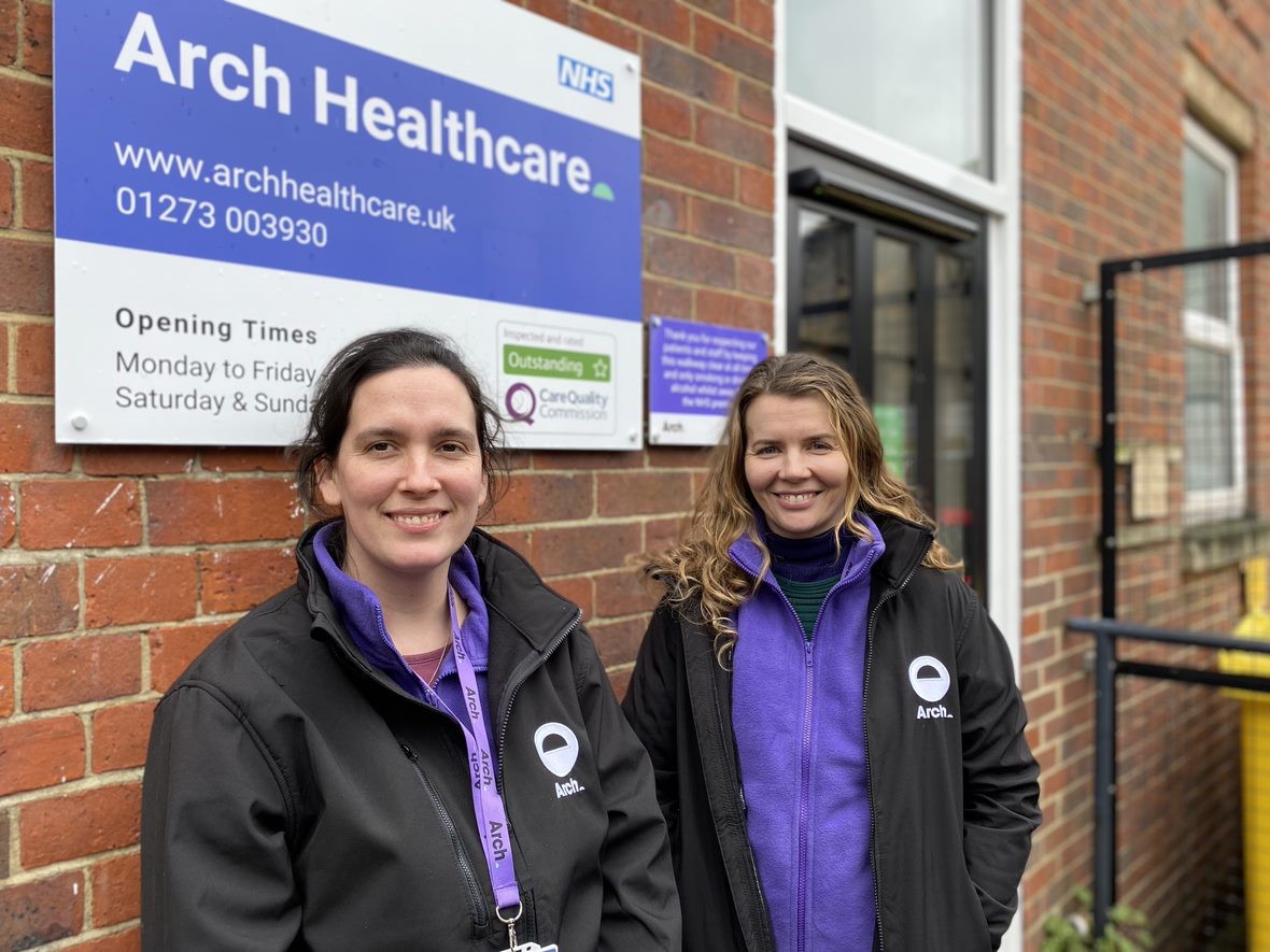 2 members of the Arch outreach team standing outside the surgery, under the sign by the front door. Smiling at the camera