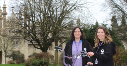 Two women standing in a park next to a bike, smiling at the camera